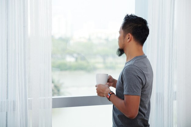 Asiatischer Mann, der vor hohem Fenster mit Becher steht und heraus schaut