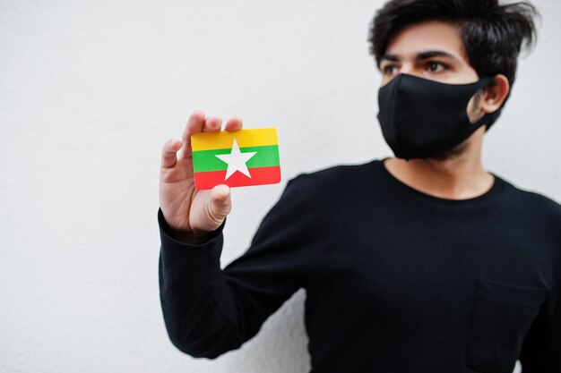 Asiatischer Mann trägt ganz schwarz mit Gesichtsmaske und hält die Flagge Myanmars in der Hand, isoliert auf weißem Hintergrund Coronavirus Country Concept