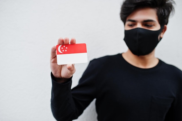 Asiatischer Mann trägt ganz schwarz mit Gesichtsmaske und hält die Flagge Singapurs in der Hand, isoliert auf weißem Hintergrund Coronavirus Country Concept