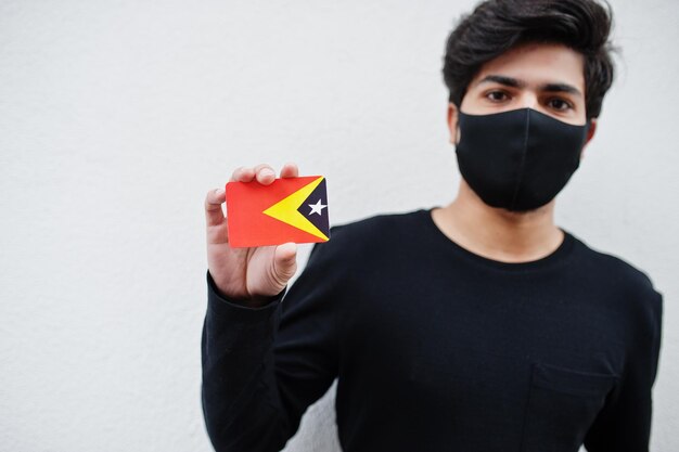 Asiatischer Mann trägt ganz schwarz mit Gesichtsmaske und hält die Osttimor-Flagge in der Hand, isoliert auf weißem Hintergrund Coronavirus Country Concept