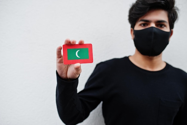 Asiatischer Mann trägt ganz schwarz mit Gesichtsmaske und hält Malediven-Flagge in der Hand, isoliert auf weißem Hintergrund Coronavirus Country Concept