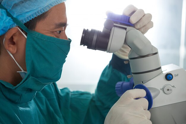 Asiatischer medizinischer Forscher im Schutzblech, in Maske, in Hut und in Handschuhen, die in Mikroskop blicken