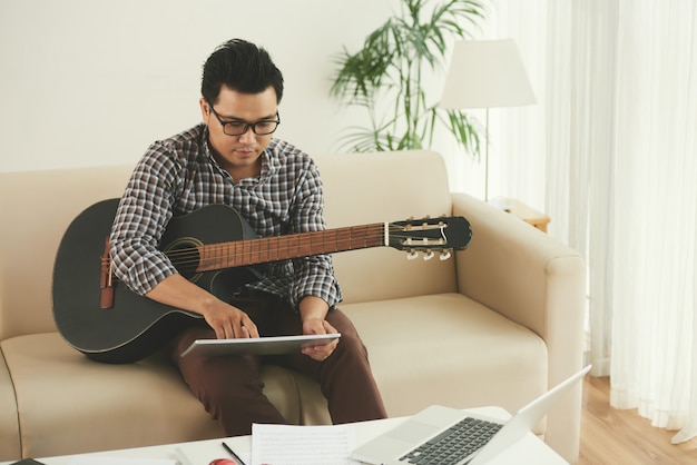 Asiatischer Musiker, der zu Hause auf Couch mit Gitarre sitzt und Tablette verwendet