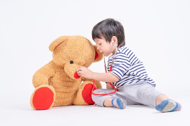 Asiatischer netter Junge, der ein Doktorgebrauchsstethoskop überprüft den großen Teddybären sitzt auf Boden spielt