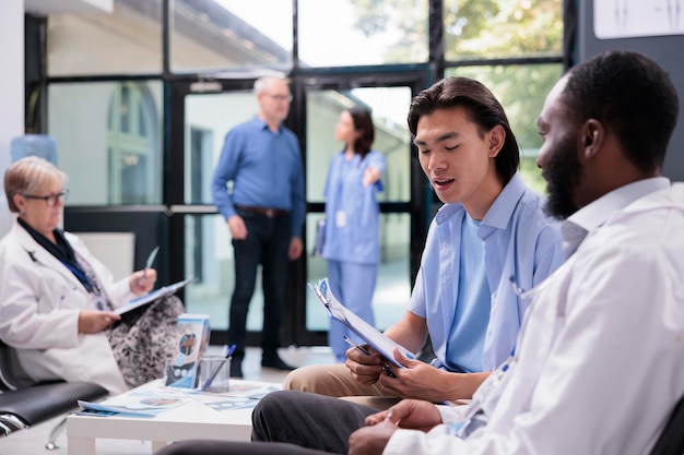 Asiatischer Patient hält Klemmbrett in der Hand und füllt medizinischen Bericht aus, während er während der Konsultation im Wartebereich des Krankenhauses mit einem Facharzt über die Gesundheitsfürsorge spricht. Verschiedene Menschen stehen an der Rezeption