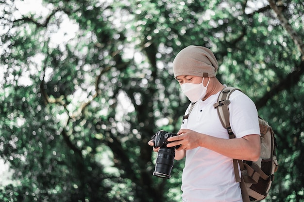 Asiatischer Reisender trägt eine medizinische Maske mit Rucksack und überprüft ein Bild vor der Kamera im Park mit Kopierbereich Reisefotograf Berufungs- und Urlaubskonzept