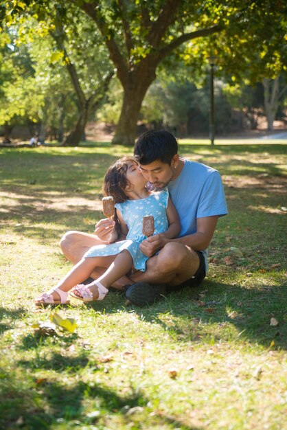 Asiatischer Vater und Tochter ruhen mit Eis im Park. Glücklicher Mann, der auf dem Boden sitzt, mit einem netten Mädchen auf den Knien, das Eis hält und ein Kind, das seine Wange küsst. Glücksmomente, Vaterschaftskonzept