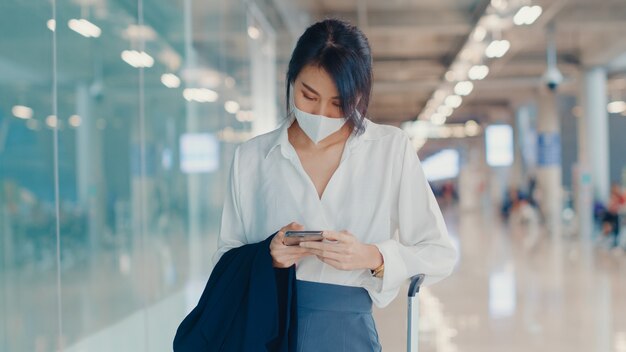 asiatisches Geschäftsmädchen, das Smartphone für Scheck-Bordkarte verwendet, die mit Gepäck zum Terminal am Inlandsflug am Flughafen geht.