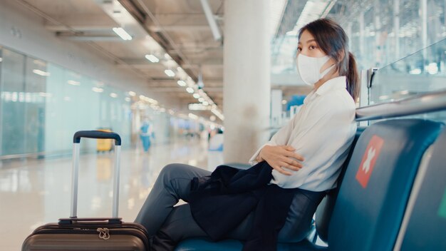 asiatisches Geschäftsmädchen mit Gepäck, das in der Bank sitzt, warten und Partner für Flug am Flughafen suchen.