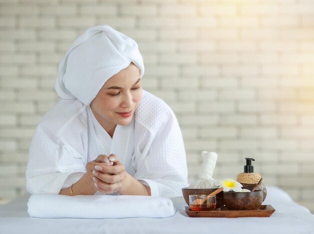 Asiatisches glückliches, entspanntes weibliches modell in weißem, sauberem bademantel legte sich auf ein massagebett mit kompressenkugel-kokosöl und plumeria-blume in einem tablett, das mit dem smartphone lächelte und auf eine körpermassage-therapie wartete.