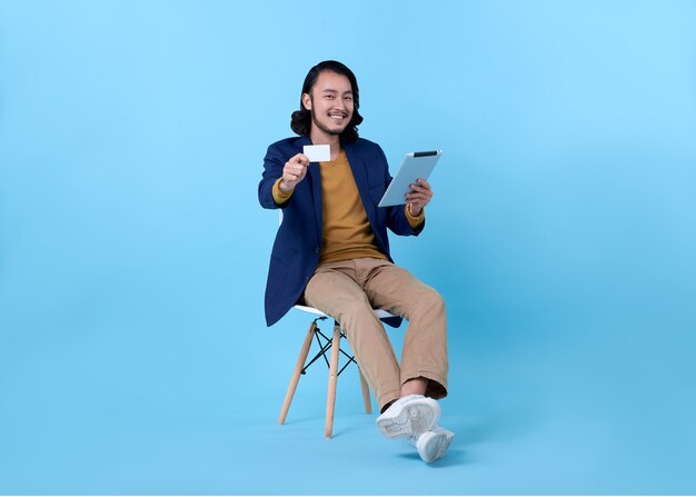 Asiatisches glückliches Lächeln des Geschäftsmannes, das Kreditkarte zeigt und eine digitale Tablette verwendet, während auf Stuhl auf hellem Blau sitzt.