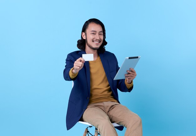 Asiatisches glückliches Lächeln des Geschäftsmannes, das Kreditkarte zeigt und eine digitale Tablette verwendet, während auf Stuhl auf hellem Blau sitzt.