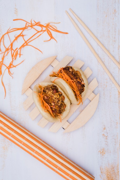 Asiatisches Gua Bao diente auf kreisförmiger hölzerner Platte mit Essstäbchen und zerriebener Karotte auf Holzoberfläche