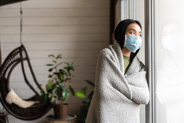 Kostenloses Foto asiatisches mädchen, das krank wird und eine medizinische gesichtsmaske trägt, die in der covid-quarantäne bleibt und nach draußen schaut, ja...