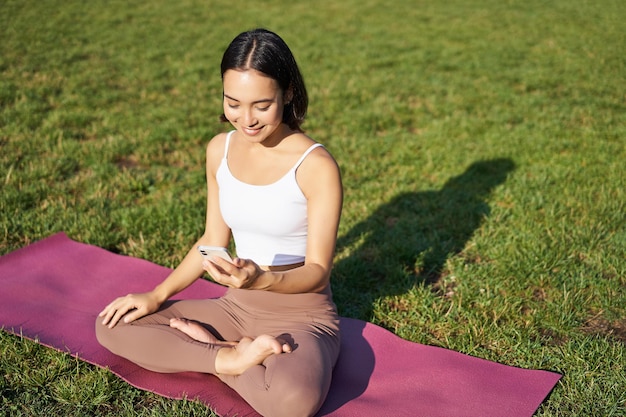 Asiatisches Mädchen folgt der Yoga-Trainings-App, schaut auf das Smartphone, meditiert und macht frische Übungen