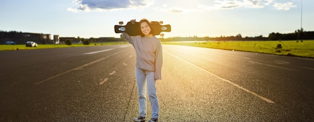 Kostenloses Foto asiatisches mädchen mit skateboard, das während des sonnenuntergangs auf der straße steht, posieren mit ihrem langen board-cruiser