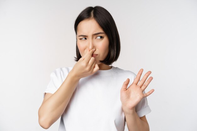 Asiatisches Mädchen sieht angewidert aus, wenn es Produkte mit schlechtem Geruch ablehnt, die Nase vor Abneigung schließen und vor weißem Hintergrund stehen