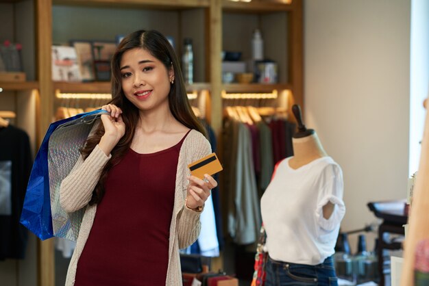 Asiatisches Mädchen, welches die Plastikkarte und Einkaufstasche stehen im Kleidungsspeicher hält