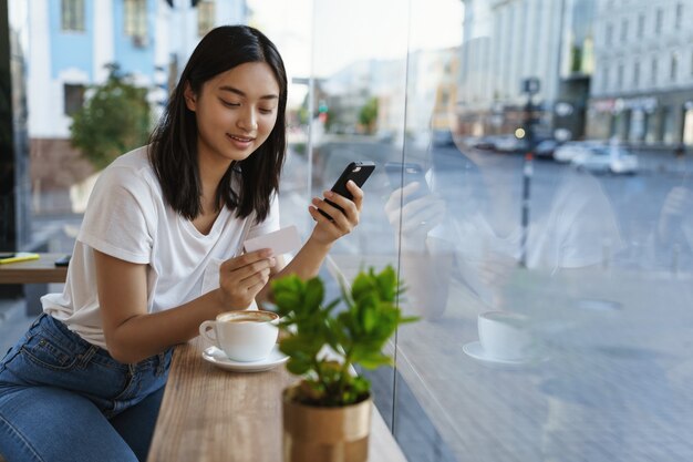 Asiatisches modernes Mädchen in einem Café, Kaffee trinken, gibt die Kreditkartennummer in Smartphone ein.