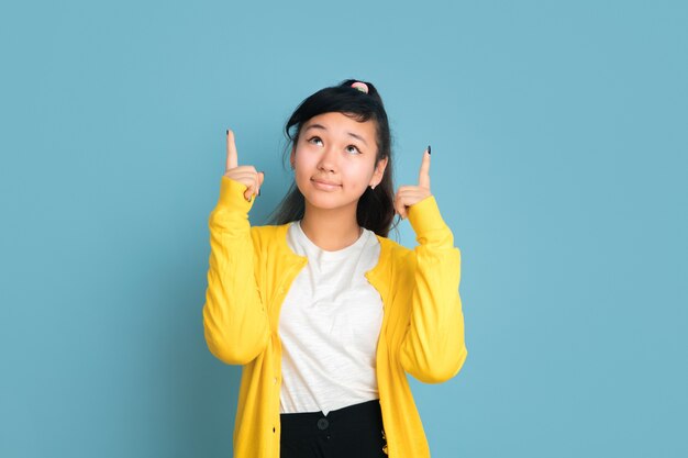 Asiatisches Teenagerporträt lokalisiert auf blauem Studiohintergrund. Schönes weibliches brünettes Modell mit langen Haaren im lässigen Stil. Konzept der menschlichen Emotionen, Gesichtsausdruck, Verkauf, Anzeige. Nach oben zeigend.