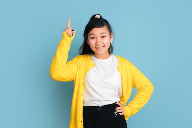 Asiatisches Teenagerporträt lokalisiert auf blauem Studiohintergrund. Schönes weibliches brünettes Modell mit langen Haaren. Konzept der menschlichen Emotionen, Gesichtsausdruck, Verkauf, Anzeige. Lächeln, nach oben zeigen, sieht süß aus.