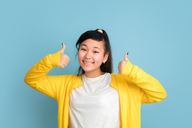 Asiatisches Teenagerporträt lokalisiert auf blauem Studiohintergrund. Schönes weibliches brünettes Modell mit langen Haaren. Konzept der menschlichen Emotionen, Gesichtsausdruck, Verkauf, Anzeige. Lächelnd, Daumen hoch, zeigt.