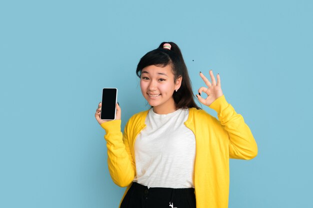 Asiatisches Teenagerporträt lokalisiert auf blauem Studiohintergrund. Schönes weibliches brünettes Modell mit langen Haaren. Konzept der menschlichen Emotionen, Gesichtsausdruck, Verkauf, Anzeige. Leeren Telefonbildschirm anzeigen.