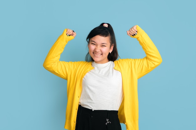 Asiatisches Teenagerporträt lokalisiert auf blauem Studiohintergrund. Schönes weibliches brünettes Modell mit langen Haaren. Konzept der menschlichen Emotionen, Gesichtsausdruck, Verkauf, Anzeige. Viel Spaß beim Gewinnen, Wettkonzept.