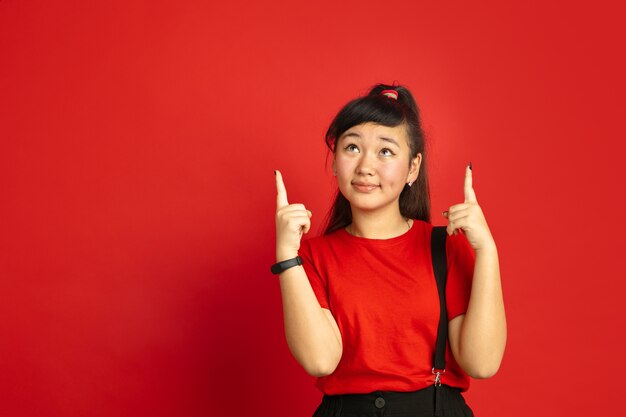 Asiatisches Teenagerporträt lokalisiert auf rotem Studiohintergrund. Schönes weibliches brünettes Modell mit langen Haaren im lässigen Stil. Konzept der menschlichen Emotionen, Gesichtsausdruck, Verkauf, Anzeige. Nach oben zeigend.