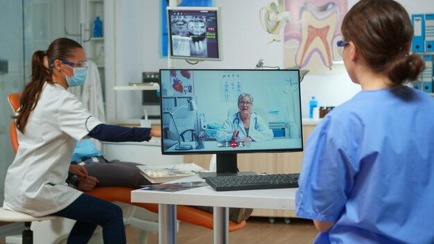 Assistent, der einen Videoanruf mit einem erfahrenen stomatologischen Arzt unter Verwendung des Computers hat, während der Arzt mit dem Patienten im Hintergrund arbeitet. Krankenschwester hört Zahnarzt auf Webcam auf Stuhl im stomatologischen Büro sitzend