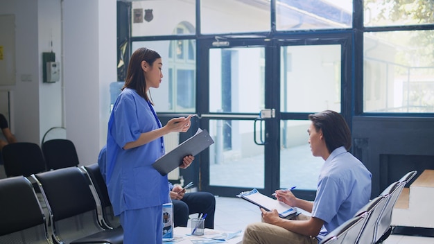 Kostenloses Foto assistent steht am krankenhaus-schalter und schreibt medizinische expertise auf die schreibtafel. ein asiatischer patient ruft die krankenschwester an, um ihm während des kontrollbesuchs im wartezimmer mit versicherungsinformationen zu helfen