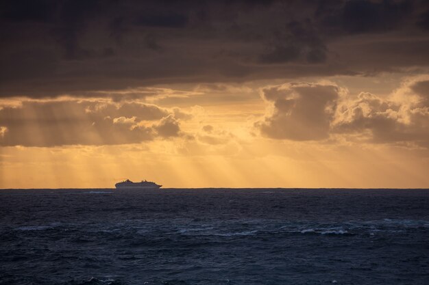 Atemberaubende Aufnahme des ruhigen blauen Ozeans und der Silhouette eines Schiffes unter einem bewölkten Himmel während des Sonnenuntergangs