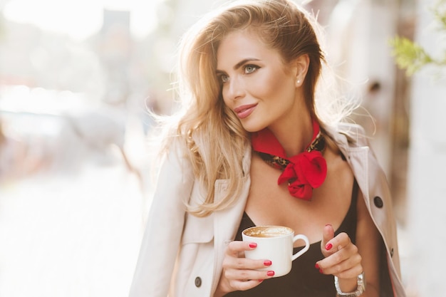 Atemberaubende blonde Dame mit rotem Halstuch, die einen guten Tag genießt und leckeren Latte trinkt. Outdoor-Porträt einer fröhlichen modischen Frau trägt eine beige Jacke und hält eine Tasse Kaffee.
