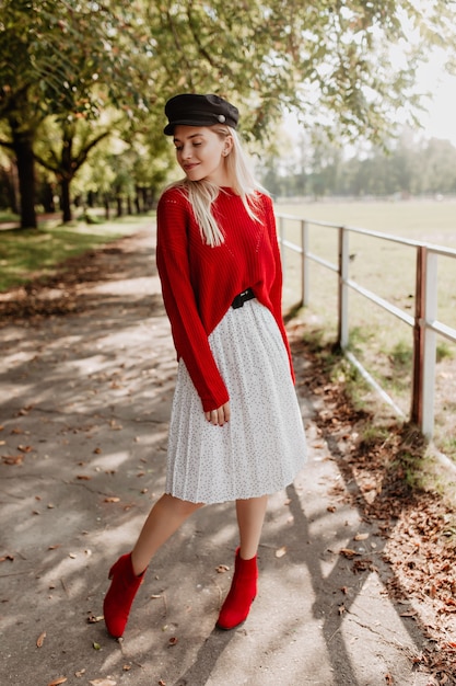 Atemberaubende blondine mit trendigem roten pullover und stilvollem kleid mit dunklem, schönem hut. schönes modell, das souverän unter abgefallenen blättern aufwirft.