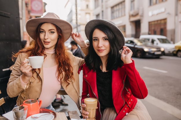 Atemberaubende brünette Frau im grauen Fedora, die Zeit mit Ingwerfreundin im Café verbringt