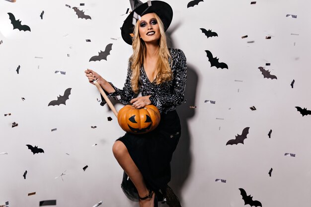 Atemberaubende Hexe mit hellem Make-up, das Halloween-Kürbis hält. Glamouröse blonde Frau in gruseliger Kleidung, die sich auf Horrorparty vorbereitet.