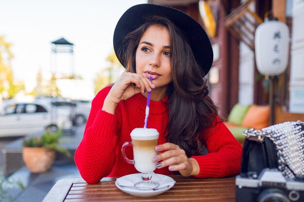 Atemberaubende junge Dame in stilvollem schwarzen Hut und leuchtend rotem Pullover, der im offenen Raumcafé sitzt und Kaffee mit Milch oder Cappuccino trinkt.