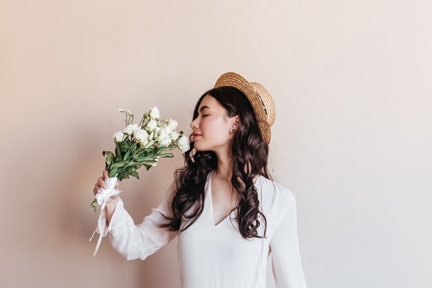 Atemberaubende lockige asiatische Frau, die weiße Blumen schnüffelt. Studioaufnahme der romantischen chinesischen Dame mit Eustomas.