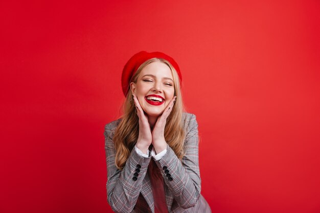 Atemberaubendes französisches Mädchen, das auf roter Wand aufwirft. Frohe blonde junge Frau lächelnd
