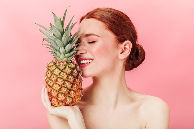 Atemberaubendes Ingwermädchen, das Ananas mit geschlossenen Augen hält Studioaufnahme einer wunderschönen kaukasischen Frau mit Früchten, die auf rosa Hintergrund posiert