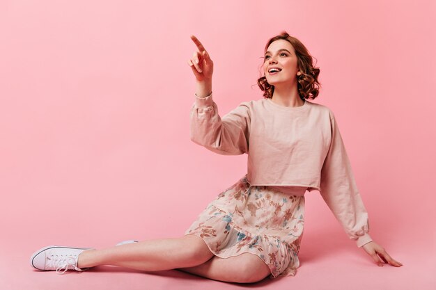 Atemberaubendes Mädchen im Rock, das auf rosa Hintergrund mit Lächeln sitzt. Inspirierte lockige junge Frau, die mit dem Finger zeigt.