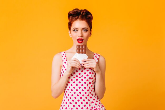 Atemberaubendes Pinup-Mädchen mit roten Lippen, die Schokolade essen. Vorderansicht der jungen Frau des Ingwers im gepunkteten Kleid lokalisiert auf gelbem Raum.