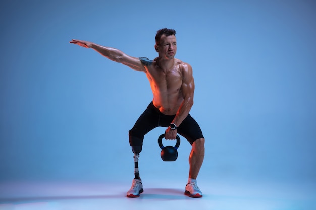 Athlet mit Behinderungen oder Amputierter lokalisiert auf blauem Studiohintergrund. Professioneller männlicher Sportler mit Beinprothesentraining mit Gewichten in Neon.