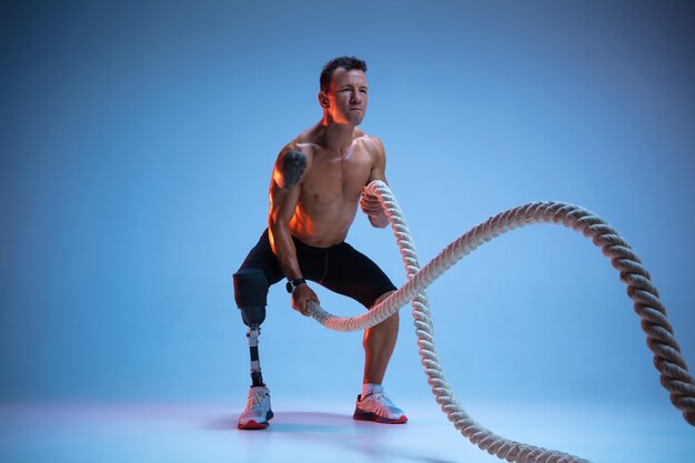 Athlet mit Behinderungen oder Amputierter lokalisiert auf blauem Studiohintergrund. Professioneller männlicher Sportler mit Beinprothesentraining mit Gewichten in Neon.
