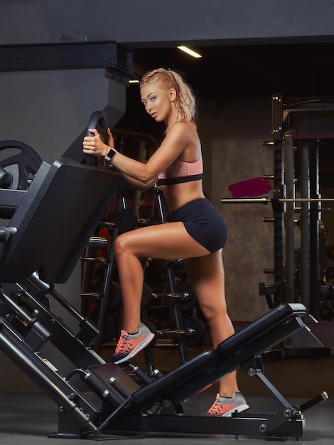 Athletische blonde Frau in Sportkleidung posiert in der Nähe der Beinpresse in einem Fitnessstudio.
