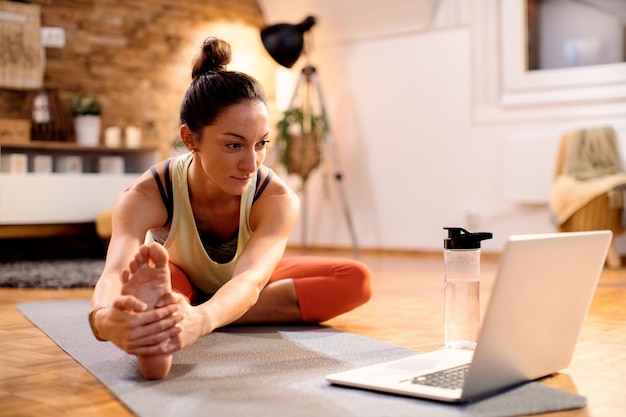 Athletische Frau, die sich auf dem Boden streckt, während sie einen Laptop benutzt und Anweisungen im Internet befolgt