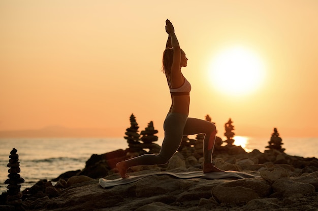 Athletische Frau mit der Hand über dem Kopf, die in der Abenddämmerung Yoga-Balance-Übungen auf einem Strandfelsen macht.