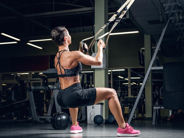 Athletische Frau mittleren Alters mit kurzen Haaren, die in einem Fitnessstudio mit Trx-Aufhängungsstreifen trainiert.