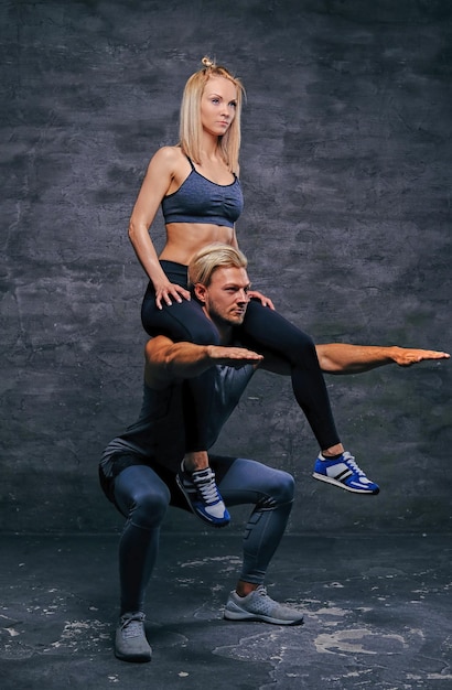 Athletischer blonder Mann, der Kniebeugen mit schmal geschnittener Frau auf seinen Schultern macht.