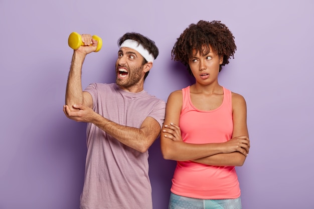 Kostenloses Foto athletischer mann hebt hantel, hat müdes training, trägt stirnband und t-shirt, unglückliche gelangweilte frau steht mit verschränkten armen